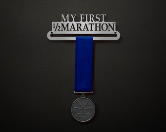 Könnte beinhalten: Ein silberner Medaillenhalter mit dem Text "MY FIRST 1/2 MARATHON" und einem blauen Band, das daran hängt. Eine silberne Medaille mit einem Läufermotiv hängt am Band.