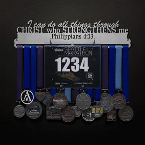 May include: A metal medal display rack with the inscription "I can do all things through CHRIST who STRENGTHENS me Philippians 4:13". The rack is holding a variety of medals, including a blue and silver medal with the text "Seattle Marathon" and the number "1234".