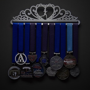 Op de afbeelding: Een zilveren medailleschaal met een decoratieve hartvormige bovenkant en een silhouet van een hardlopende figuur. De schaal heeft acht haken en houdt een verscheidenheid aan medailles vast met blauwe, paarse en lichtblauwe linten. De medailles hebben verschillende ontwerpen, waaronder een cirkel met de letter A, een rood-wit-blauw schild, een zwart-wit kompas, een zilveren hardlopende figuur, een rood-wit logo met de tekst "Rosarville Express" en een verscheidenheid aan andere ontwerpen.