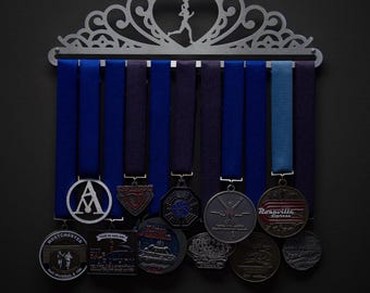 Puede incluir: Un soporte para medallas de plata con una parte superior decorativa en forma de corazón y una silueta de una figura corriendo. El soporte tiene ocho ganchos y sostiene una variedad de medallas con cintas azules, moradas y azul claro. Las medallas presentan varios diseños, incluyendo un círculo con la letra A, un escudo rojo, blanco y azul, una brújula en blanco y negro, una figura plateada corriendo, un logotipo rojo y blanco con el texto "Rosarville Express" y una variedad de otros diseños.