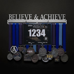 Może przedstawiać: Metalowy stojak na medale z napisem "BELIEVE & ACHIEVE" nad kilkoma medalami i wstążkami. Na wyświetlaczu znajduje się numer startowy z numerem 1234. Wstążki są niebieskie i fioletowe, a medale srebrne.