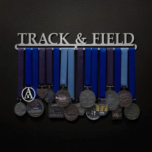 May include: A silver metal medal display rack with the words "TRACK & FIELD" on it. The rack has 12 hooks for hanging medals. There are several medals hanging on the rack, including a silver medal with the words "Roseville Express" on it.