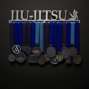 May include: A metal Jiu-Jitsu medal display rack with a silhouette of a person practicing Jiu-Jitsu. The rack has 12 hooks for hanging medals and ribbons. The rack is silver and has the word "Jiu-Jitsu" written on it. There are several medals hanging on the rack, including a silver medal with the letter "A" on it, a silver medal with the words "Ragsdale Express" on it, and a silver medal with a silhouette of a person doing a Jiu-Jitsu move on it.
