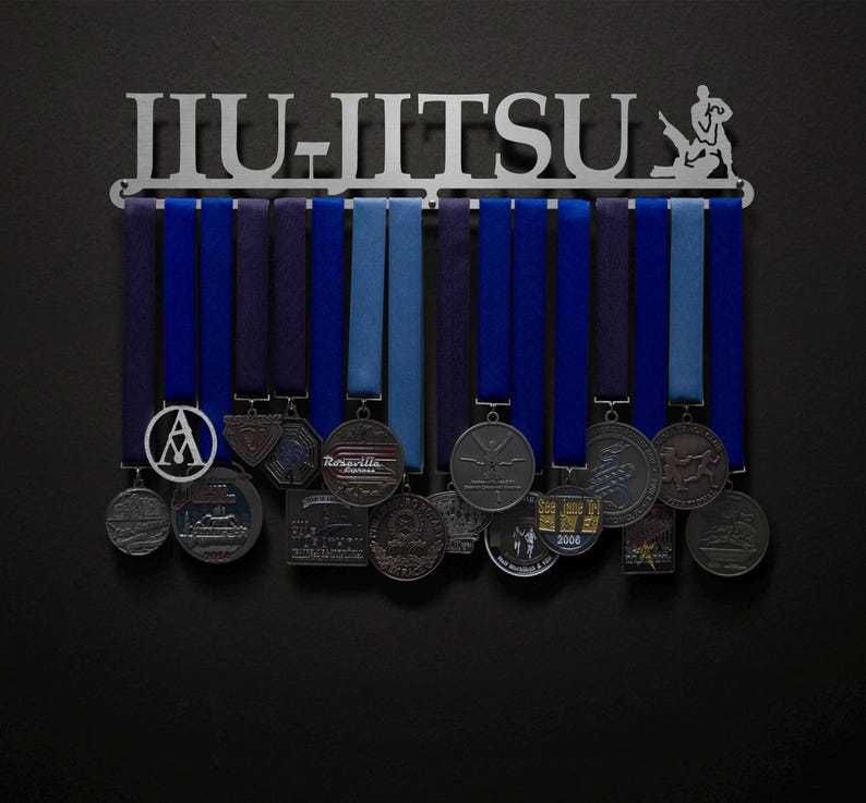 May include: A metal Jiu-Jitsu medal display rack with a silhouette of a person practicing Jiu-Jitsu. The rack has 12 hooks for hanging medals and ribbons. The rack is silver and has the word "Jiu-Jitsu" written on it. There are several medals hanging on the rack, including a silver medal with the letter "A" on it, a silver medal with the words "Ragsdale Express" on it, and a silver medal with a silhouette of a person doing a Jiu-Jitsu move on it.
