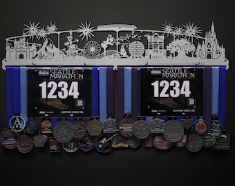 May include: A white metal medal display rack with a silhouette of a city skyline. The rack has two rows of hooks for hanging medals and two spaces for displaying race bibs. The bibs are from the Seattle Marathon and show the number 1234. The rack is decorated with several medals.