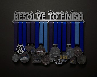 May include: A silver medal rack with the words "Courage to Start Strength to Endure RESOLVE TO FINISH" hanging on a black wall. The rack is filled with blue and purple ribbons and silver medals.