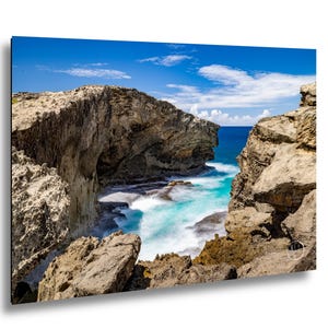 May include: A landscape photograph of a rugged, rocky coastline with a deep blue ocean inlet. The scene features textured, light brown cliffs framing turquoise water with white sea foam, under a bright blue sky with scattered white clouds.
