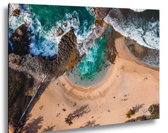 Può includere: Una vista aerea di una spiaggia sabbiosa a forma di mezzaluna che incontra l'acqua turchese dell'oceano. Formazioni rocciose scure e frastagliate costeggiano la riva, con schiuma bianca che si infrange sui bordi. Piccole figure sono visibili.