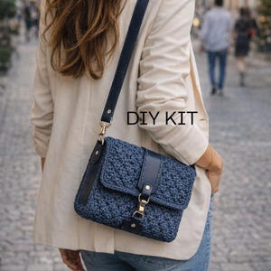 Puede incluir: Un bolso bandolera de crochet azul marino con correa y detalles de cuero. El bolso tiene un cierre de solapa con un cierre dorado. El texto "DIY KIT" es visible en la imagen.