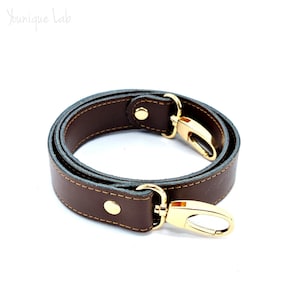 Peut inclure: Une sangle en cuir marron avec des ferrures dorées et des coutures. La sangle est pliée en cercle et possède deux fermoirs pivotants.