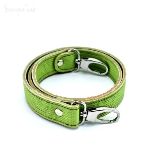 Può includere: Un cinturino per borsa verde lime arrotolato, con chiusure girevoli in metallo color argento. Il cinturino è realizzato in materiale strutturato e presenta un lato inferiore marrone chiaro. Il marchio "Younique Lab" è nell'angolo in alto a sinistra.