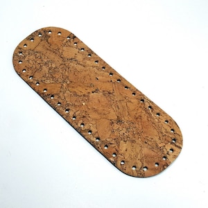 Op de afbeelding: Een rechthoekige kurken basis met afgeronde uiteinden, met een natuurlijke bruine kleur en een getextureerd oppervlak. De basis heeft gelijkmatig verdeelde gaten rondom de omtrek, ontworpen voor knutselprojecten. Op een witte achtergrond.