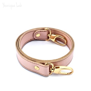 Può includere: Un cinturino in pelle rosa chiaro con accessori dorati e due fibbie girevoli. Il cinturino è piegato in un cerchio.