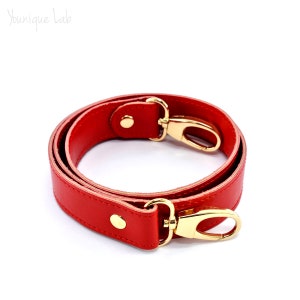 Peut inclure: Sangle d'épaule en cuir rouge avec des ferrures dorées et un fermoir pivotant. La sangle est pliée en cercle.