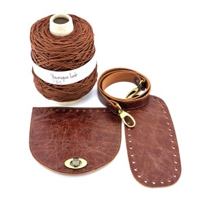 Op de afbeelding: Een bruine leren tasset met een bruine leren riem, een bruine leren klep en een spoel met bruin koord. De set bevat een metalen sluiting en een label met de tekst "Younique Lab <B13".