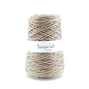 May include: A spool of beige yarn with a tightly woven texture. The label reads "Younique Lab ER2" in black script. The yarn is wound in a neat, symmetrical pattern, suitable for various crafting projects.
