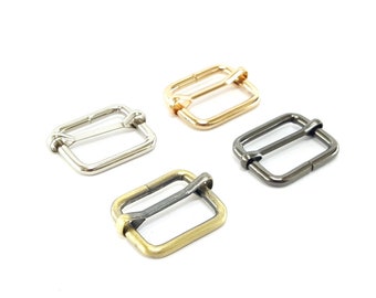 Op de afbeelding: Vier metalen schuifgespen in verschillende afwerkingen: zilver, goud, antiek messing en zwart. De gespen zijn rechthoekig met een kleine lus aan één kant.