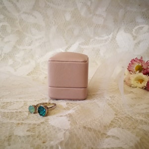 May include: A pink ring box with a silver ring with two turquoise gemstones. The ring box and ring are on a white lace background.
