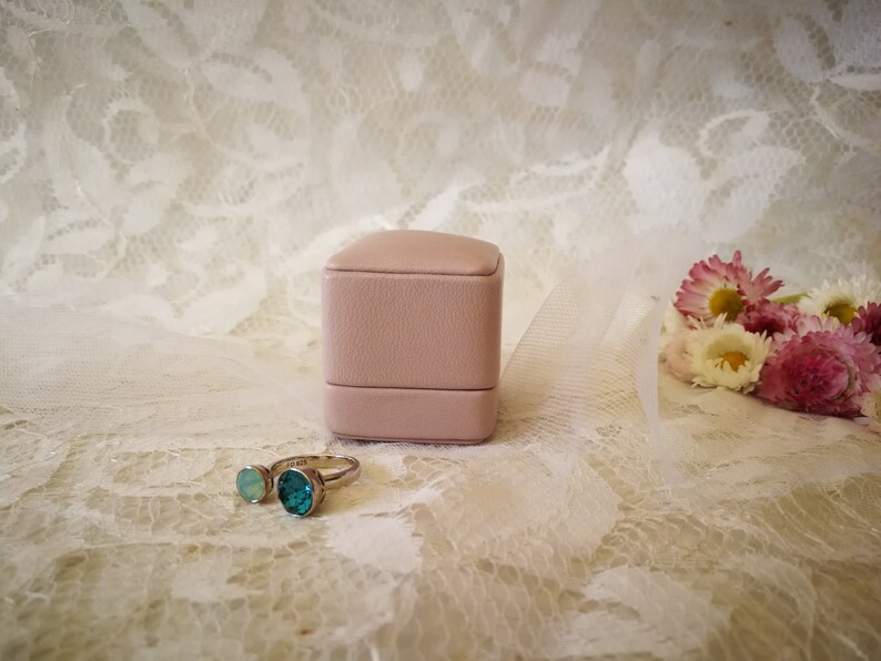 May include: A pink ring box with a silver ring with two turquoise gemstones. The ring box and ring are on a white lace background.