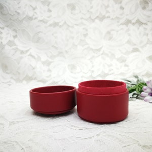 May include: A red velvet ring box with a lid and a base. The box is open and the lid is sitting next to the base. The box is on a white lace background.