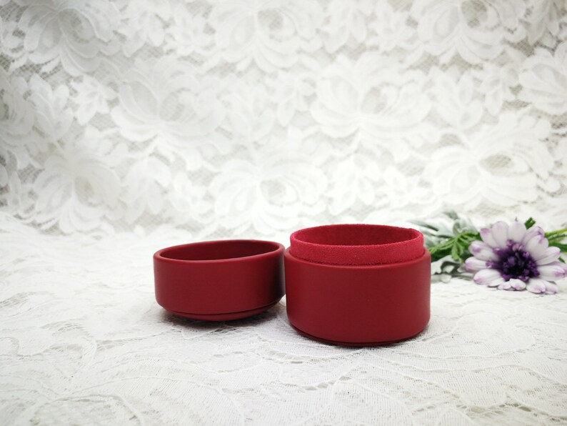 May include: A red velvet ring box with a lid and a base. The box is open and the lid is sitting next to the base. The box is on a white lace background.