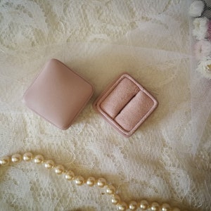 May include: Two pink ring boxes on a white lace background with a pearl necklace.
