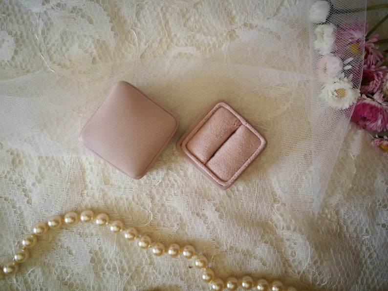 May include: Two pink ring boxes on a white lace background with a pearl necklace.
