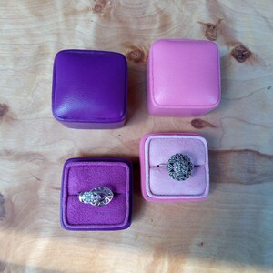 May include: Four square ring boxes in purple and pink, two closed and two open, revealing rings. One ring has a floral design, the other has an eye-like design. The boxes sit on a light wood surface.