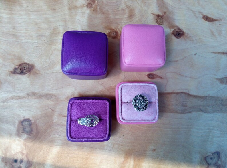 May include: Four square ring boxes in purple and pink, two closed and two open, revealing rings. One ring has a floral design, the other has an eye-like design. The boxes sit on a light wood surface.