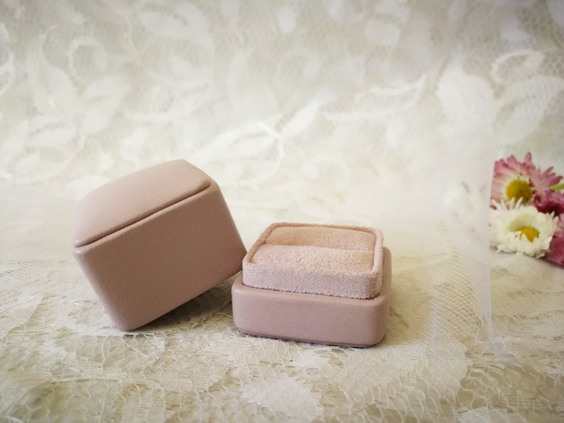 May include: Two pink ring boxes with a velvet interior. The boxes are on a white lace background.