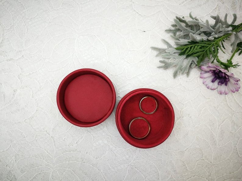 May include: Two red velvet ring boxes, one open with two gold rings inside, the other closed. The boxes are on a white lace background.