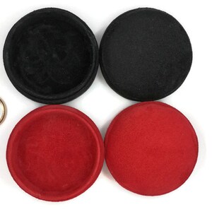 May include: An overhead shot shows four round, fabric-covered containers in black and red, along with two gold-colored metal rings. The containers are open and closed, revealing the interior. The background is white.