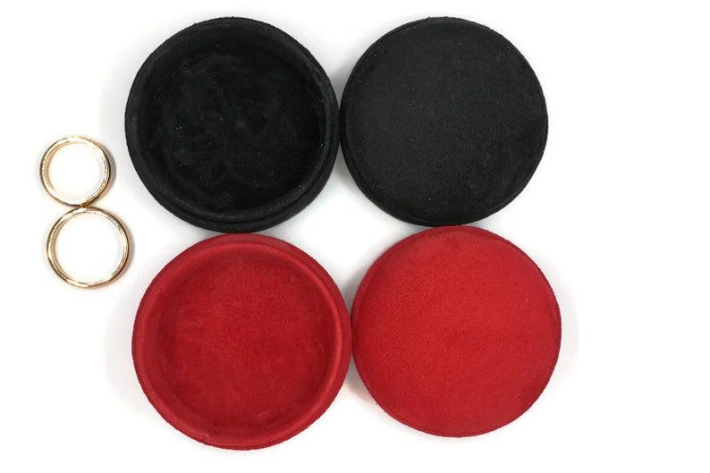 May include: An overhead shot shows four round, fabric-covered containers in black and red, along with two gold-colored metal rings. The containers are open and closed, revealing the interior. The background is white.