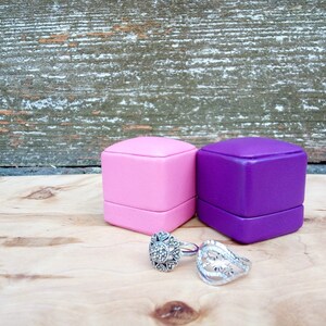 May include: Two ring boxes, one pink and one purple, sit on a wooden surface next to two silver rings. The rings have intricate designs, one with a floral pattern and the other with a cluster of small stones. The background is a weathered wooden wall.