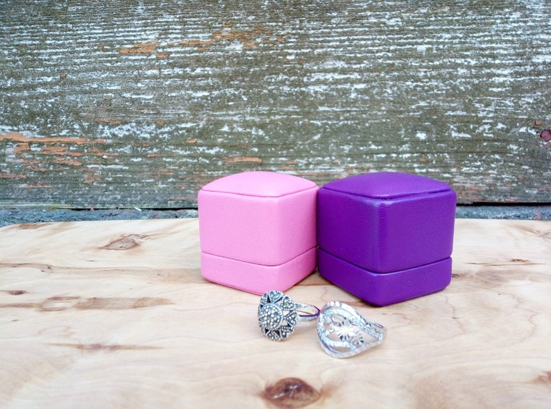 May include: Two ring boxes, one pink and one purple, sit on a wooden surface next to two silver rings. The rings have intricate designs, one with a floral pattern and the other with a cluster of small stones. The background is a weathered wooden wall.