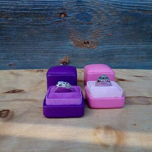 May include: Four ring boxes, two purple and two pink, each with a velvet interior. Two rings are displayed, one in each color of box. The rings have intricate designs and are set against the velvet.