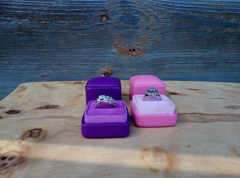 May include: Four ring boxes, two purple and two pink, each with a velvet interior. Two rings are displayed, one in each color of box. The rings have intricate designs and are set against the velvet.