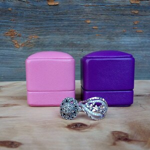 May include: Two rings and two jewelry boxes are displayed on a wooden surface. One ring features a floral design with clear stones, and the other has a dark stone center. The boxes are pink and purple.