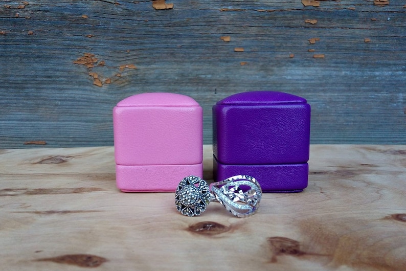 May include: Two rings and two jewelry boxes are displayed on a wooden surface. One ring features a floral design with clear stones, and the other has a dark stone center. The boxes are pink and purple.
