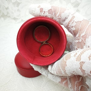 May include: Two gold wedding bands in a red velvet ring box. The box is held by a white lace glove.
