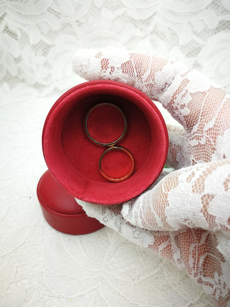 May include: Two gold wedding bands in a red velvet ring box. The box is held by a white lace glove.