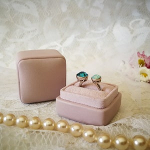 May include: Two rings with teal and green gemstones are displayed in a pink velvet ring box. The box is sitting on a white lace surface with a pearl necklace.