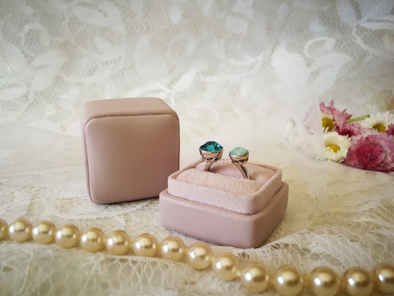May include: Two rings with teal and green gemstones are displayed in a pink velvet ring box. The box is sitting on a white lace surface with a pearl necklace.