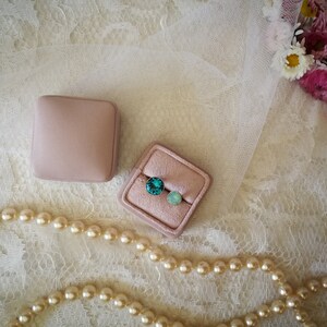 May include: Two small pink velvet boxes with turquoise and light blue gemstones inside. The boxes are on a white lace background with a pearl necklace.