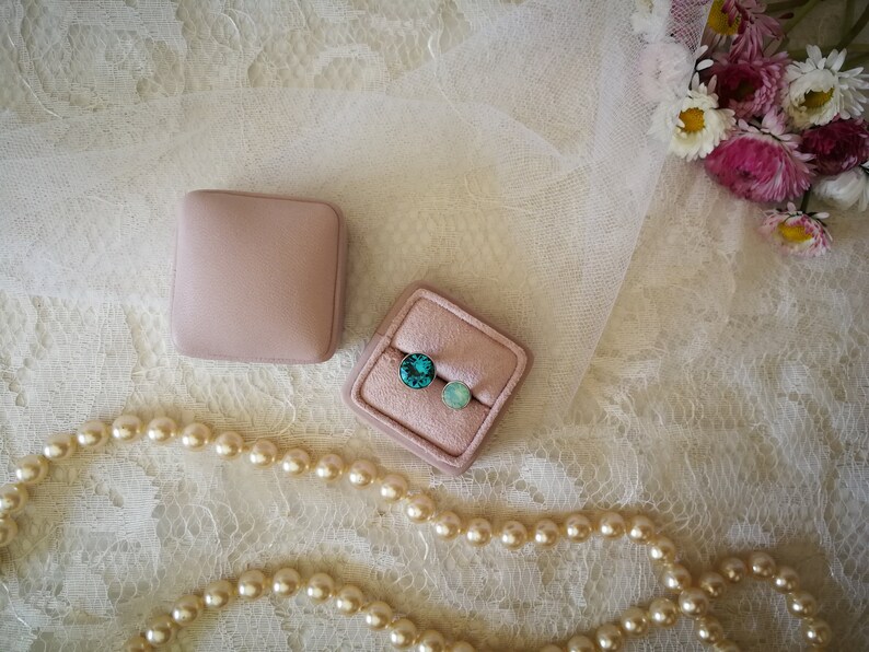May include: Two small pink velvet boxes with turquoise and light blue gemstones inside. The boxes are on a white lace background with a pearl necklace.