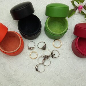 May include: Four small round leather ring boxes in black, green, red, and orange. The boxes are open and show the soft velvet lining inside. Several silver rings are scattered around the boxes.