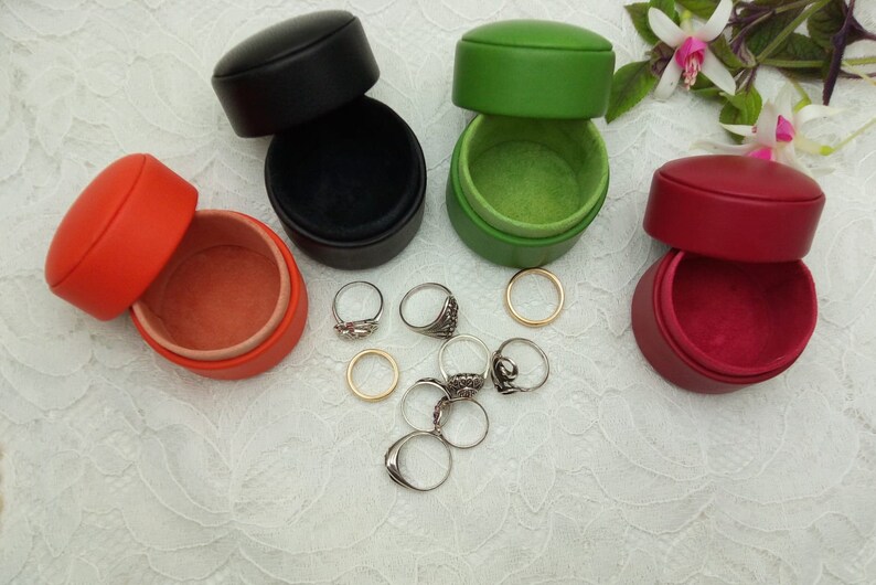 May include: Four small round leather ring boxes in black, green, red, and orange. The boxes are open and show the soft velvet lining inside. Several silver rings are scattered around the boxes.
