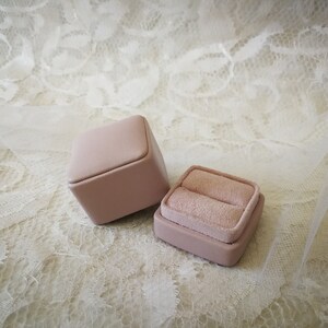 May include: Two pink ring boxes, one open and one closed, on a white lace background.