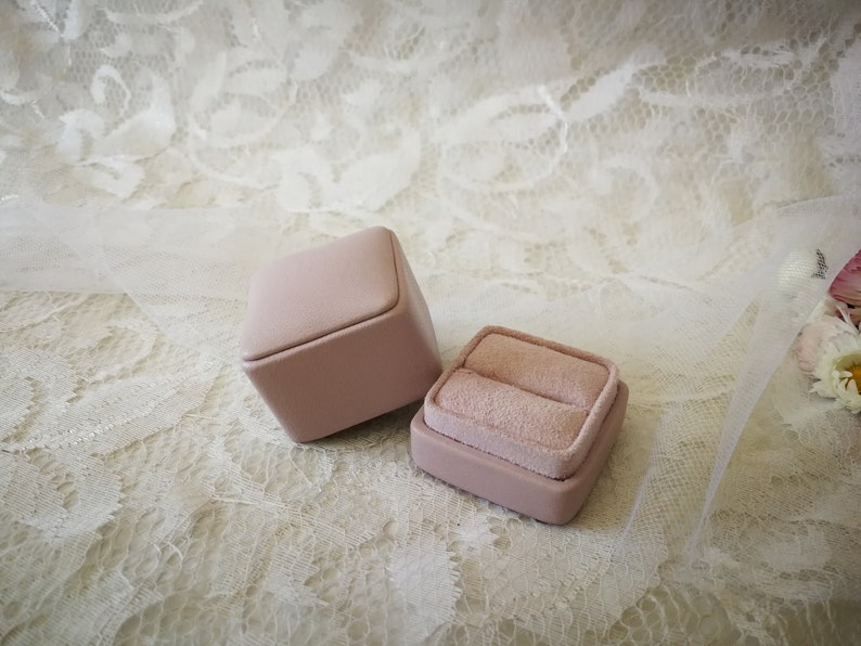 May include: Two pink ring boxes, one open and one closed, on a white lace background.