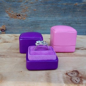 May include: A silver ring with a cluster of gemstones sits in a purple velvet-lined ring box. Two additional ring boxes, one purple and one pink, are in the background. The boxes are on a wooden surface.